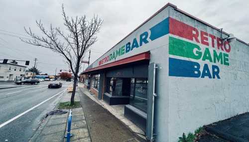 Exterior of a retro game bar with colorful signage, located on a street with overcast skies and a few passing cars.