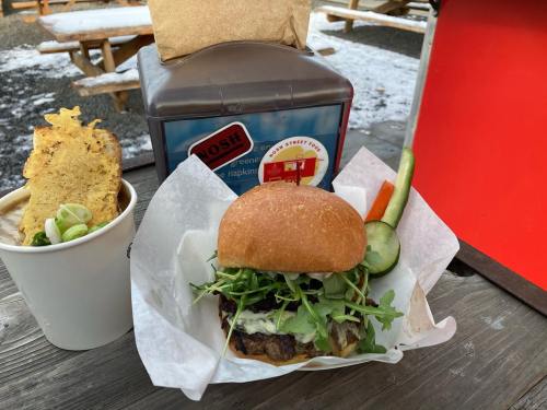 A gourmet burger with greens and sauce, served with a side of soup and vegetables, on a wooden table.