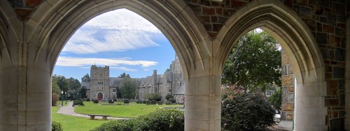 View of a stone building and green lawn framed by arched stone windows under a blue sky with clouds.