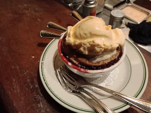 A dessert bowl with warm cobbler topped with a scoop of vanilla ice cream, surrounded by spoons on a wooden table.