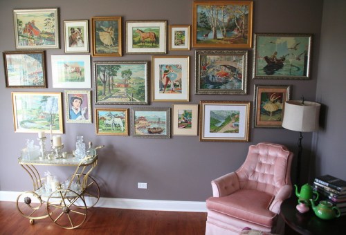 A cozy room featuring a gallery wall of framed vintage art, a pink armchair, and a decorative cart with glassware.
