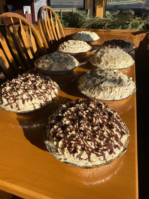 A table with various pies, including cream and chocolate toppings, displayed in a warm, sunny setting.