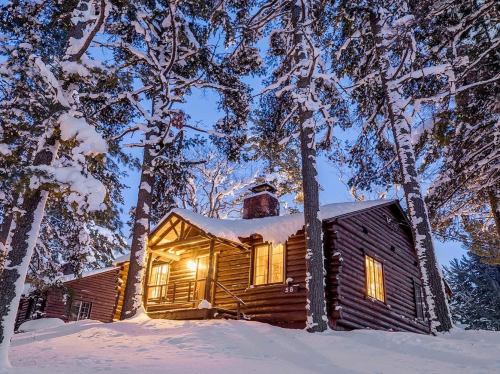 A cozy log cabin surrounded by snow-covered trees, warmly lit at dusk.