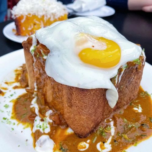 A thick slice of bread topped with a sunny-side-up egg and a rich sauce, garnished with herbs.