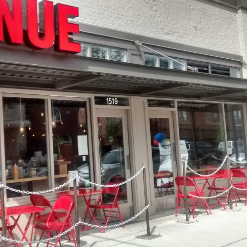 Exterior of a restaurant with red signage, featuring outdoor seating with red chairs and a chain barrier.