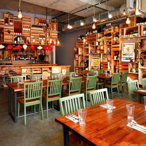 A cozy restaurant interior with wooden tables, green chairs, and shelves filled with bottles and decor. Warm lighting enhances the ambiance.