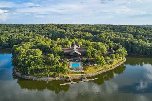 Aerial view of a modern house surrounded by trees and water, featuring a pool and a private dock.