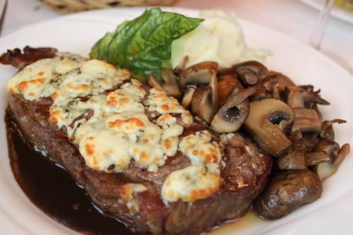 A plate featuring a cheesy steak, creamy mashed potatoes, and sautéed mushrooms, garnished with a leaf of basil.