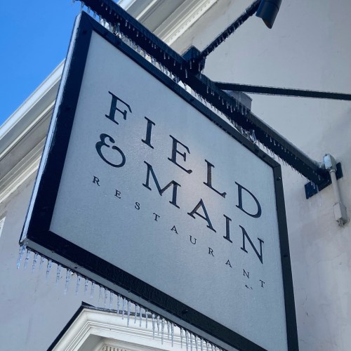 Sign for "Field & Main Restaurant" with icicles hanging from it, set against a clear blue sky.