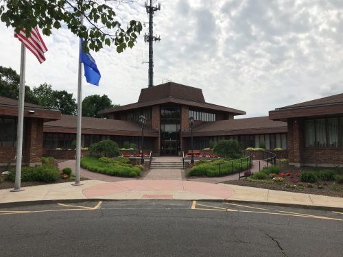 A brick building with a central entrance, surrounded by landscaped gardens and flags, under a cloudy sky.