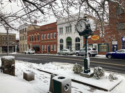 A snowy street scene featuring a vintage clock, brick buildings, and parked cars along a quiet road.