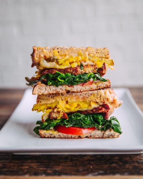 A tall sandwich stacked with layers of spinach, tomato, cheese, and yellow mustard on whole grain bread, served on a plate.