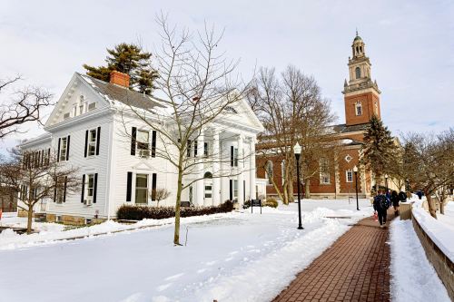 A snowy campus scene featuring a white building and a brick tower, with students walking along a path.