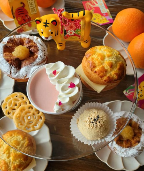 A glass platter with various pastries, a pink dessert with whipped cream, and a decorative tiger figurine, surrounded by oranges.