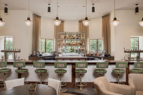 A modern bar with green stools, a well-stocked shelf of liquor, and large windows letting in natural light.