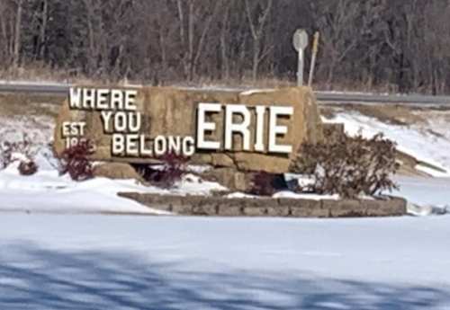Sign reading "Where You Belong Erie" with a stone base, surrounded by snow and trees in the background.