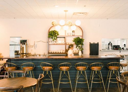 A modern café interior featuring a wooden bar, stylish stools, and plants, with a barista in motion behind the counter.