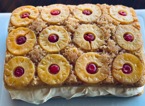 A rectangular cake topped with pineapple slices and maraschino cherries, with a creamy filling visible at the bottom.