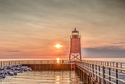 A lighthouse stands on a pier at sunset, casting a warm glow over the calm water and rocky shoreline.