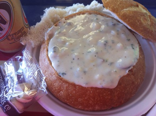 A bread bowl filled with creamy soup, accompanied by a roll and a can of cream soda.