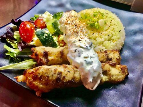 Grilled chicken skewers with rice, salad, and a creamy sauce on a black plate.