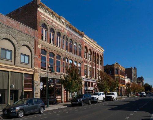 Historic brick buildings line a street under a clear blue sky, with trees and parked cars in the foreground.