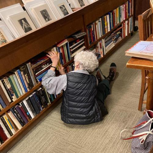 An elderly person crouches on the floor, reaching for books on a library shelf, surrounded by rows of books.