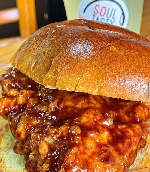 A close-up of a BBQ chicken sandwich with a glossy bun and sauce, next to a drink labeled "Soul Tasty."