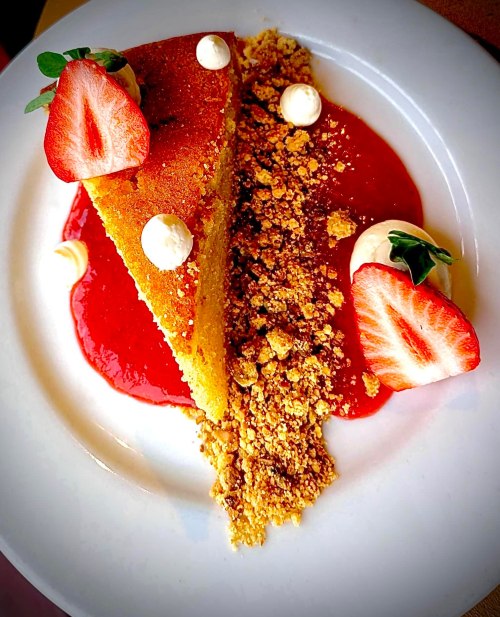 A slice of cake on a plate, garnished with strawberries, cream, and a berry sauce, with crumbled topping.