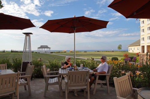 Two people sit at a patio table under umbrellas, overlooking a scenic view of the ocean and a gazebo in the distance.