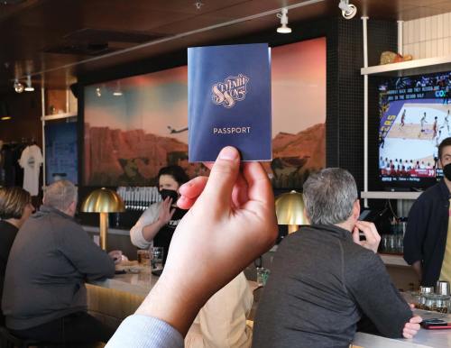 A hand holds a blue passport at a bar, with patrons and a TV showing a basketball game in the background.