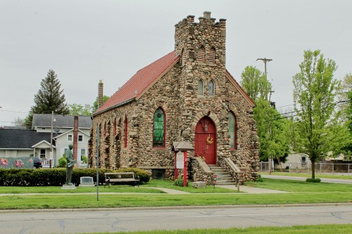 A stone church with a red roof and stained glass windows, surrounded by greenery and a small park.