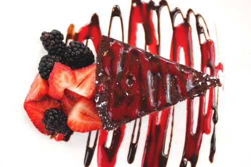 A slice of chocolate cake drizzled with berry sauce, accompanied by fresh strawberries and blackberries on a white plate.