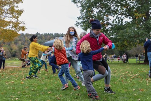 A group of children and adults dance together in a park, surrounded by trees and autumn foliage.