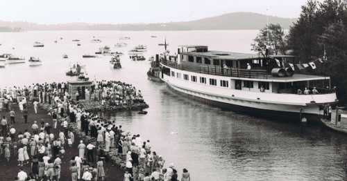 A historic black-and-white photo of a large boat arriving at a dock, with crowds gathered onshore and boats in the water.