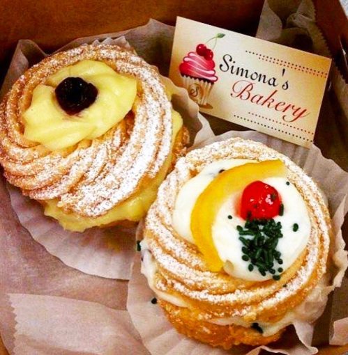 Two decorated pastries in a box, one with cream and a cherry, the other with lemon and green sprinkles, alongside a bakery card.