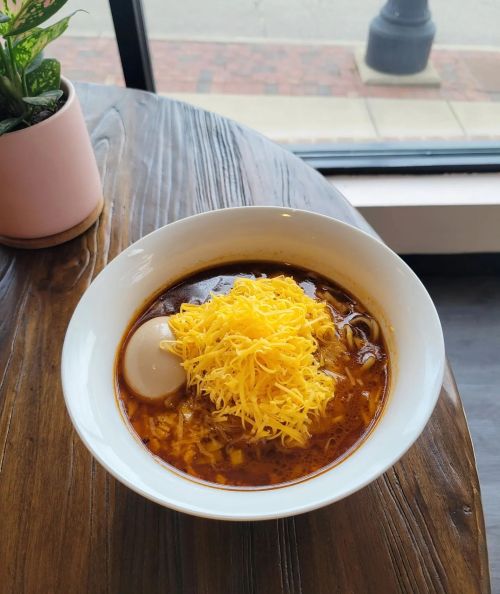 A bowl of spicy soup topped with shredded cheese and a soft-boiled egg, placed on a wooden table by a window.