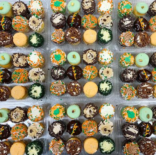 A colorful assortment of decorated cupcakes in various flavors and designs arranged in clear containers.
