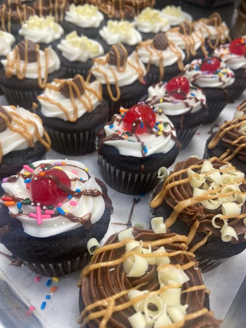 A variety of decorated chocolate cupcakes topped with frosting, cherries, sprinkles, and drizzles of caramel and chocolate.