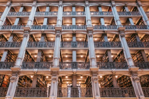 A grand library interior with multiple levels of bookshelves and ornate architectural details. Soft lighting illuminates the space.