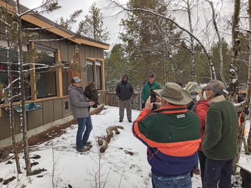 A group of people watches a man holding a bird outside in a snowy, wooded area near a building.