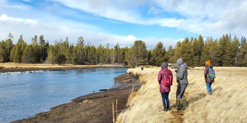 Three people walk along a riverbank surrounded by tall grass and trees under a partly cloudy sky.