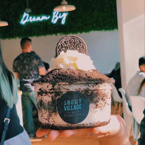 A hand holding a dessert cup topped with whipped cream and an Oreo, with a "Dream Big" neon sign in the background.