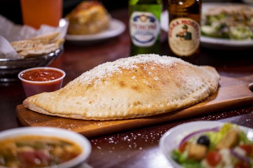 A golden-brown calzone on a wooden board, served with marinara sauce and surrounded by drinks and side dishes.