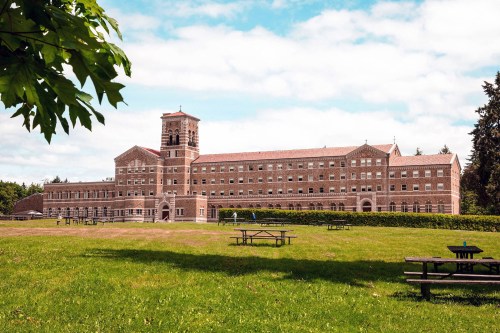 A large brick building with a tower, surrounded by green grass and trees under a partly cloudy sky.
