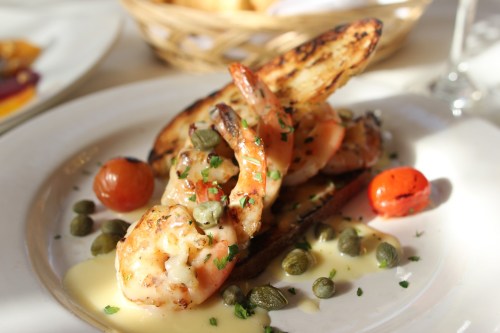 A plate of grilled shrimp on toasted bread, garnished with capers, cherry tomatoes, and a creamy sauce.