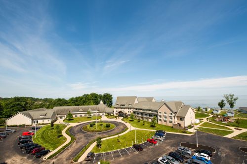 Aerial view of a hotel with a circular driveway, surrounded by green lawns and parked cars, near a body of water.