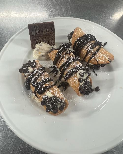 Three cannoli drizzled with chocolate sauce, topped with chocolate chips, and served with a dollop of cream on a white plate.