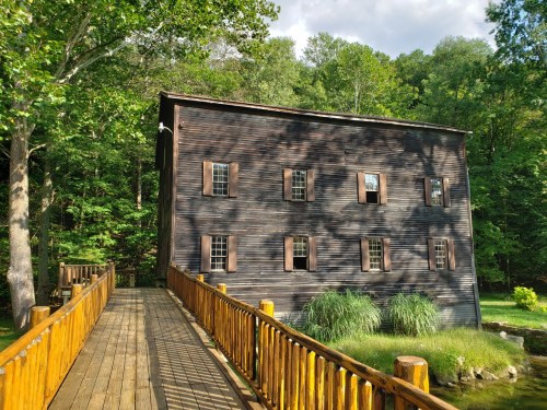 A rustic wooden building with multiple windows, surrounded by greenery, connected by a wooden bridge.