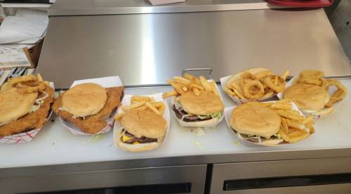 A row of burgers and fries on a counter, featuring a fried chicken sandwich and onion rings.